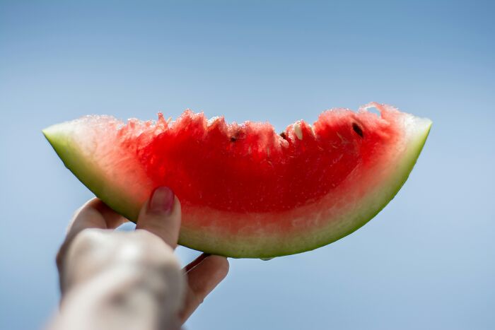 Hand holding a partially eaten watermelon slice against a clear blue sky, illustrating side effects of eating watermelon. - 1
