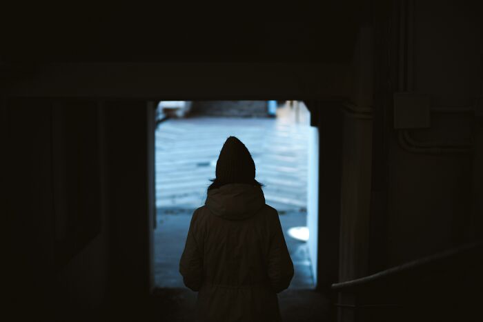 Person wearing a jacket and beanie standing alone in a dark tunnel looking towards a bright open space outside.