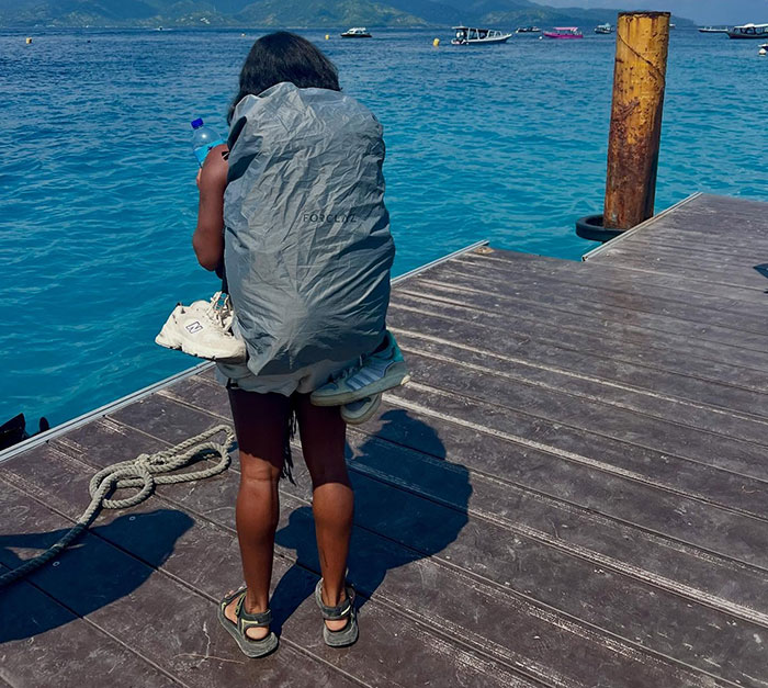 Tourist standing on a wooden dock by clear blue water, carrying hiking gear and a large backpack cover.