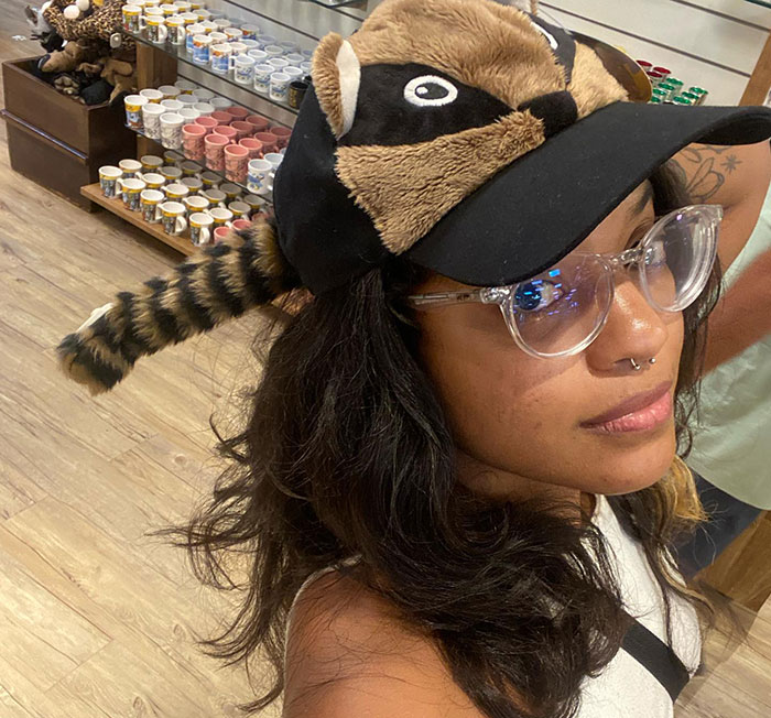 Young woman wearing clear glasses and a raccoon hat in a store with shelves of colorful mugs behind her