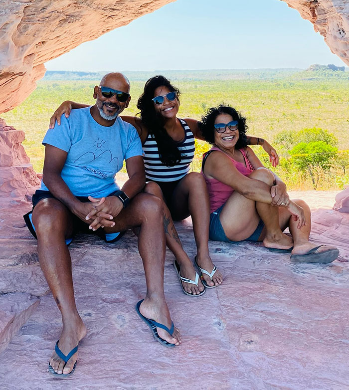 Three people wearing sunglasses sitting inside a rocky cave with a view of green landscape outside, related to hiker volcano story.