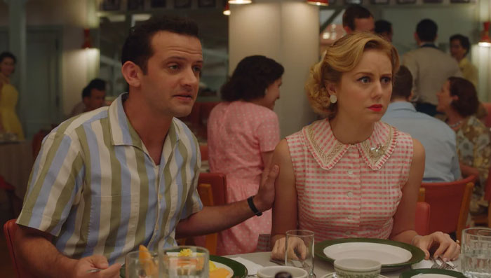 Man in striped shirt comforts Justine Lupe in pink dress at a dinner table, showcasing Justine Lupe screen performances.