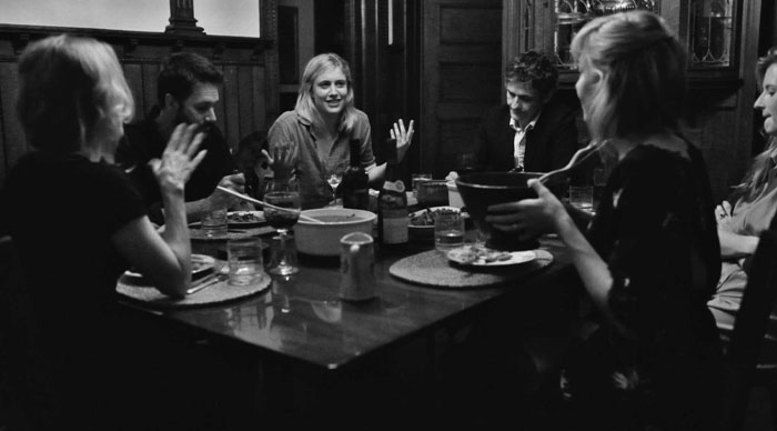 Group of people seated around a table in a dimly lit room featuring Justine Lupe in a screen performance scene.