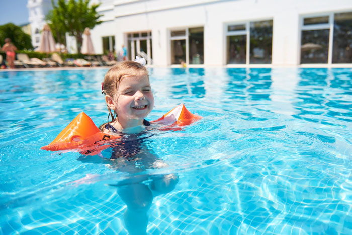 Child wearing orange floaties happily swimming in a hotel pool on a sunny day near hotel building and lounge chairs. - 9