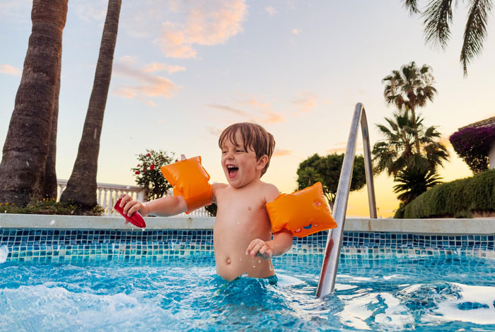 Young boy wearing floaties playing joyfully in a hotel pool at sunset near palm trees and tropical plants. - 5