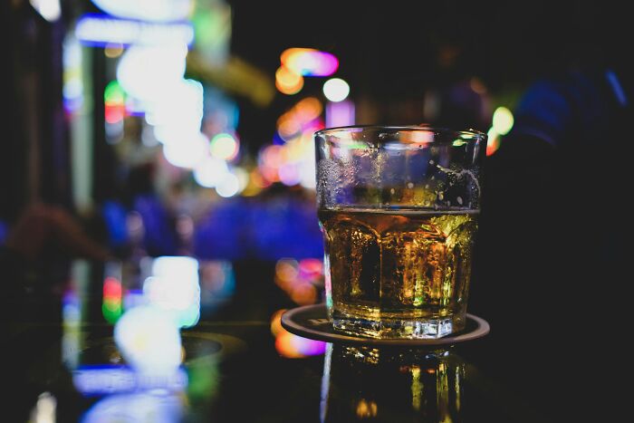 A glass of whiskey on a coaster at a dimly lit bar with colorful blurred lights in the background, showing red flag signs. - 18