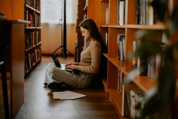 Young woman sitting on library floor using laptop focused on improving communicating calmly skills in quiet space