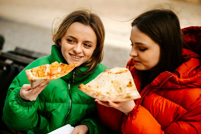 Two young women in colorful jackets eating large pizza slices outdoors, sharing bizarre facts about their countries.