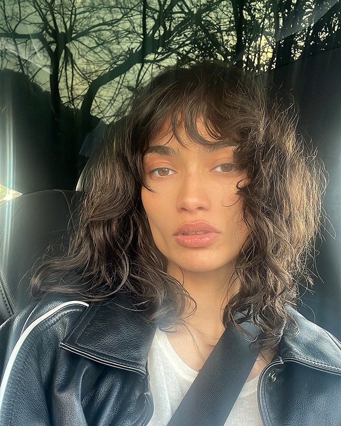 Kelly Gale with natural makeup and curly hair, wearing a leather jacket and white shirt in a car selfie.
