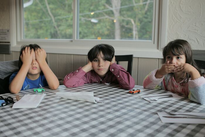Three children sitting at a checkered tablecloth, looking frustrated and bored in a home setting.
