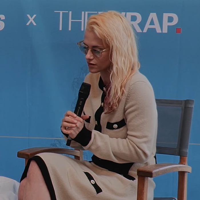 Kristen Stewart with glasses and blonde hair, holding a microphone while seated, showcasing a dramatic transformation. Kristen Stewart with glasses and blonde hair, holding a microphone while seated, showcasing a dramatic transformation.