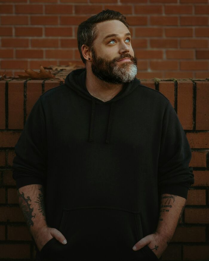 Bearded man in black hoodie with tattoos, standing against a brick wall, representing private investigator with weirdest cases. - 10