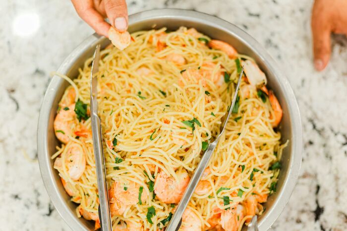 Close-up of a bowl of shrimp pasta with tongs and a hand grabbing a piece, illustrating a casual meal setting.