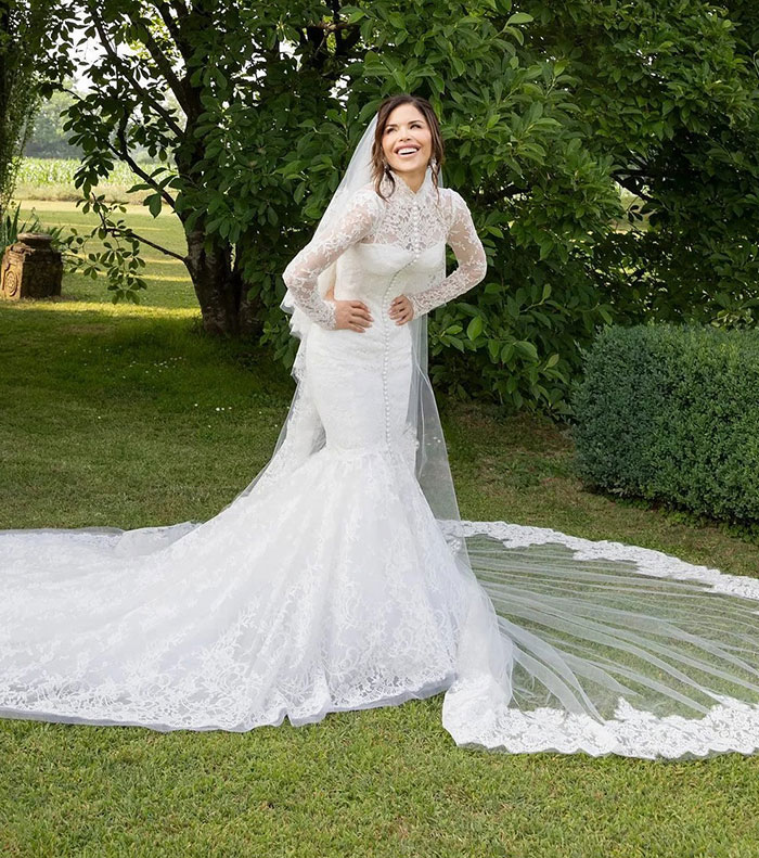 Lauren Sanchez wearing a white lace wedding dress outdoors, smiling with hands on hips, no visible diamond ring.