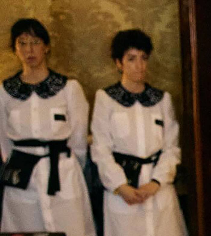 Two women in matching white outfits with black details standing against a patterned wall in a blurred photo.