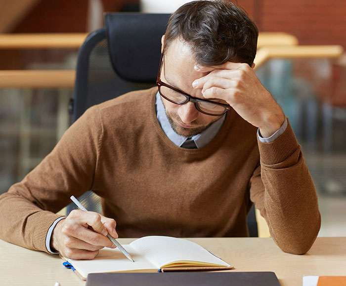Worried male teacher wearing glasses writing in notebook, highlighting literacy crisis concerns in US education setting.