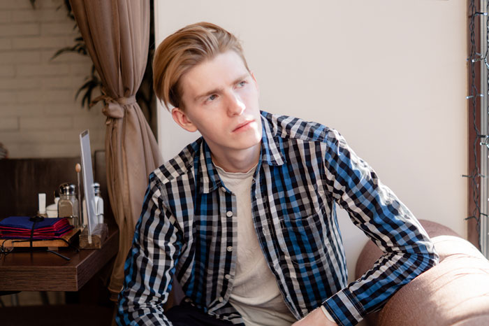 Teen sitting indoors, wearing a plaid shirt, looking thoughtful while coping with loss and working multiple jobs.