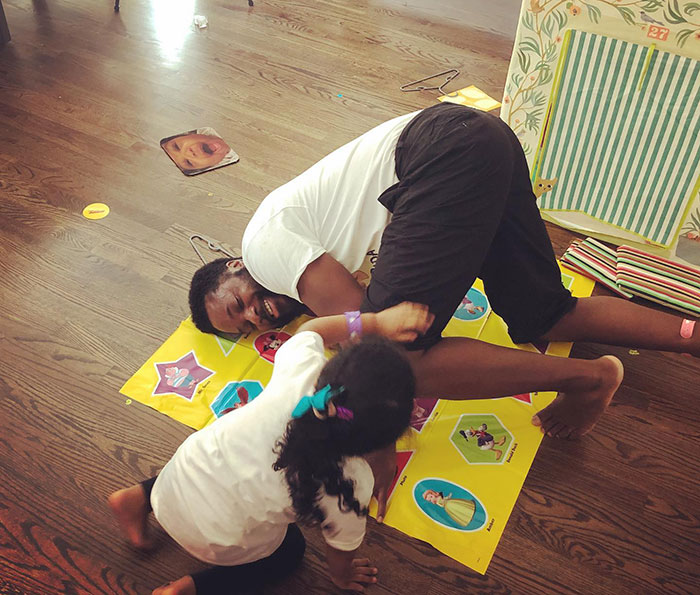 Malcolm-Jamal Warner playing a floor game and laughing with a young child during a candid moment indoors.