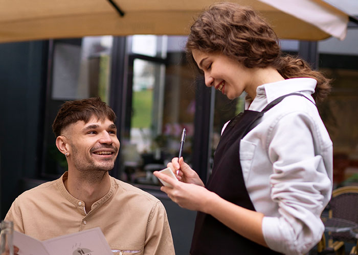 Man smiling and talking to a waitress taking an order at an outdoor caf&eacute;, capturing a casual interaction scene.