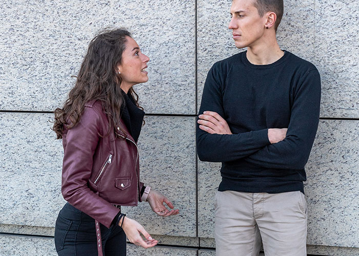 Woman with curly hair in a maroon jacket talking intensely to a man with arms crossed wearing a black sweater by a stone wall
