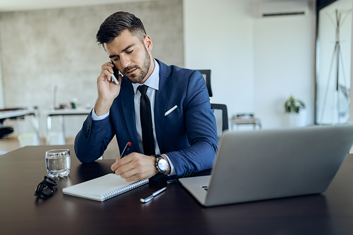 Man in a blue suit making a phone call while taking notes at a desk with a laptop and glass of water in a modern office. Man in a blue suit making a phone call while taking notes at a desk with a laptop and glass of water in a modern office.