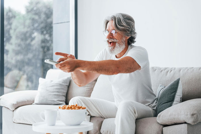Man on patio enjoying a relaxed moment, smiling and pointing with remote while sitting on couch with snacks.