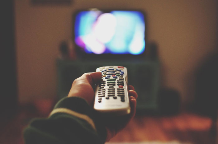 Person holding a remote control pointing towards a blurry TV screen in a dimly lit living room patio setting.