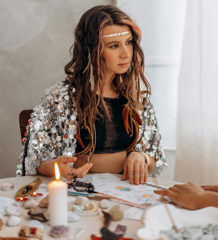 Woman with braided hair and silver shawl performing a ritual at a table, relating to sister&rsquo;s rebirth party decision.