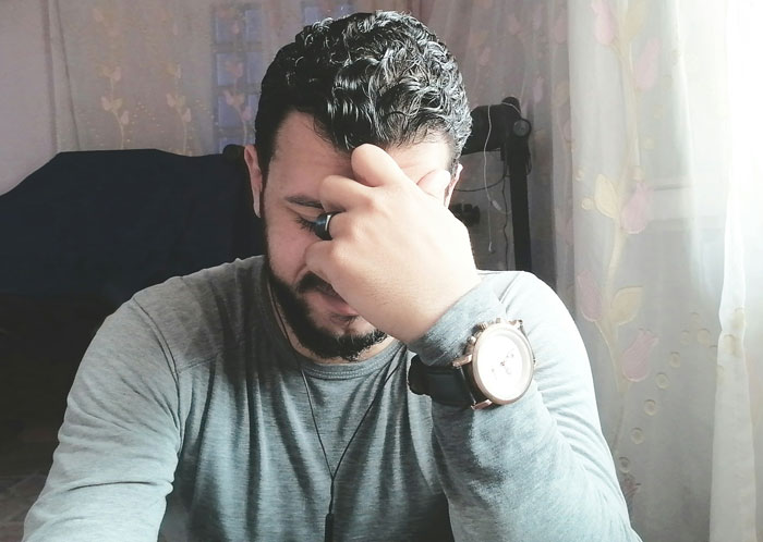 Man covering face with hand, appearing uncomfortable and contemplative, sitting indoors near a window with curtains.
