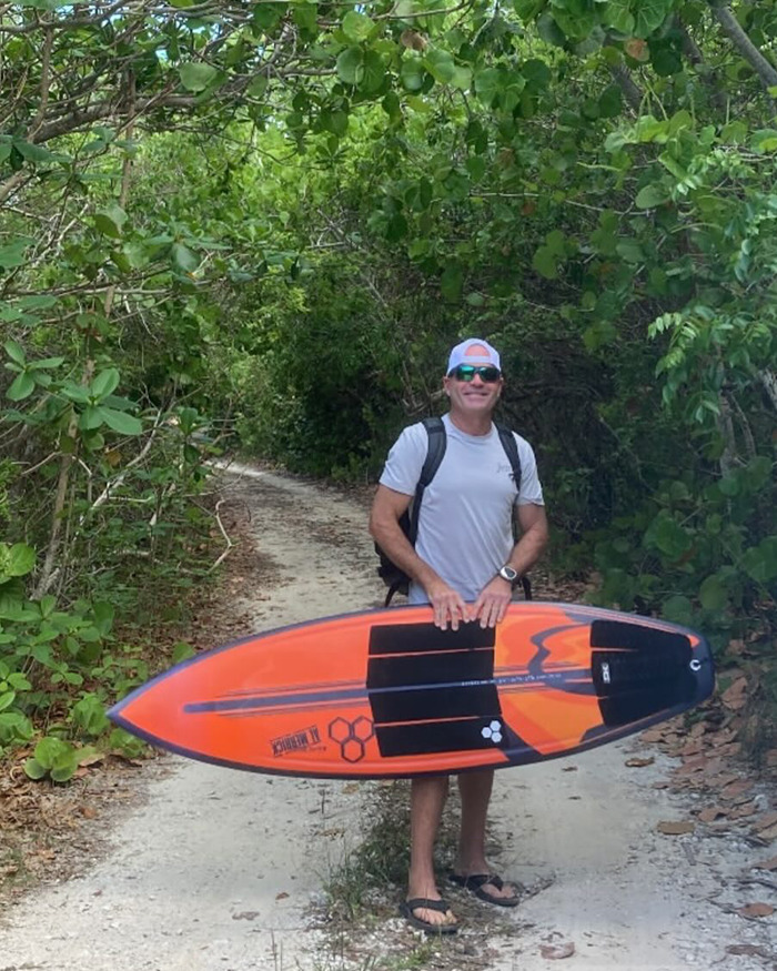 Man standing on a forest path holding an orange surfboard, representing Florida surfer involved in flying shark encounter.