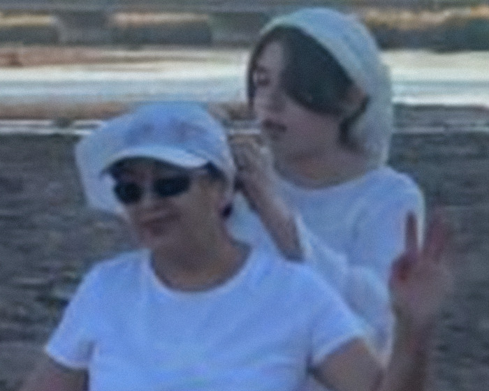 Two women outdoors near water, one fixing the other's headscarf, with calm expressions in a casual setting.