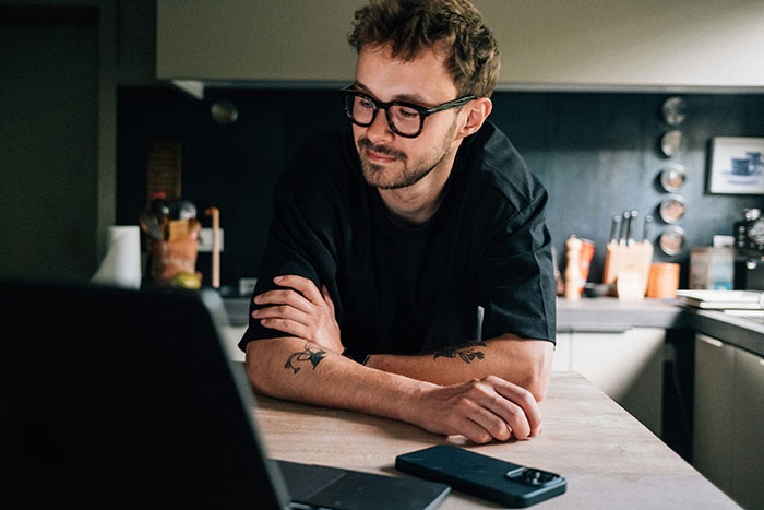 Man with glasses and tattoos sitting at kitchen table looking at laptop, illustrating father and son lock horns conflict.