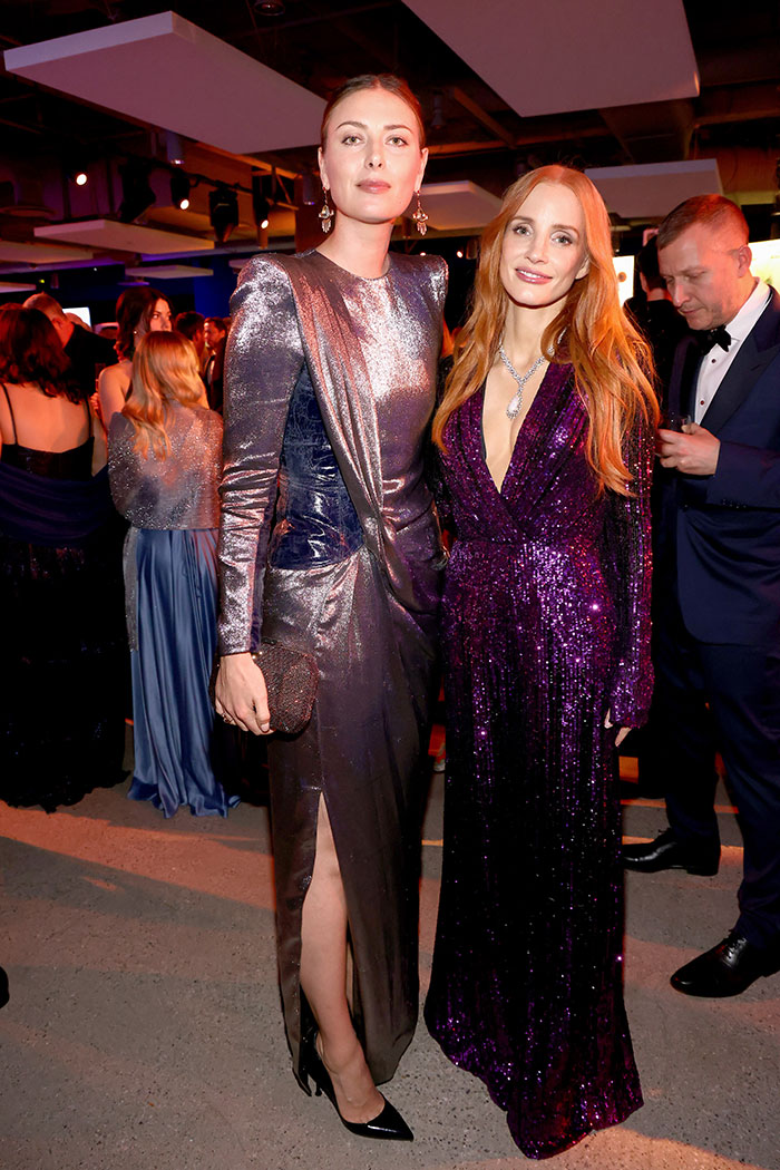 Two super tall stars dressed in metallic and sequined gowns posing at an elegant event with other guests in the background.