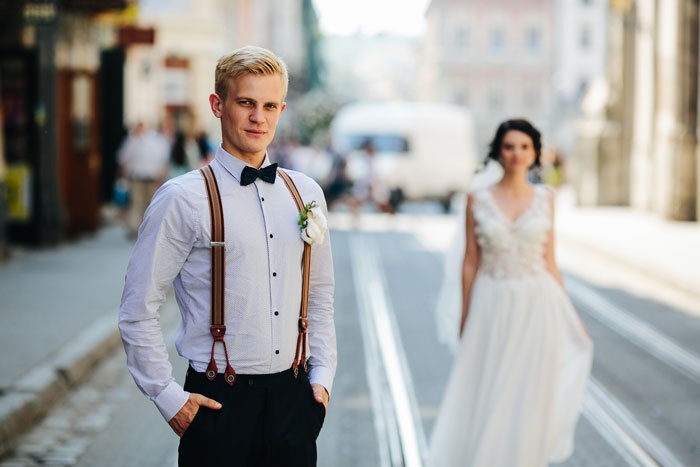 Groom standing on city street in suspenders and bow tie while bride in white wedding dress walks behind him. - 19