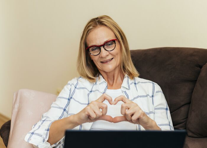 Middle-aged woman with glasses smiling and making a heart shape with hands while sitting on a couch using a laptop, related to wife's infidelity.