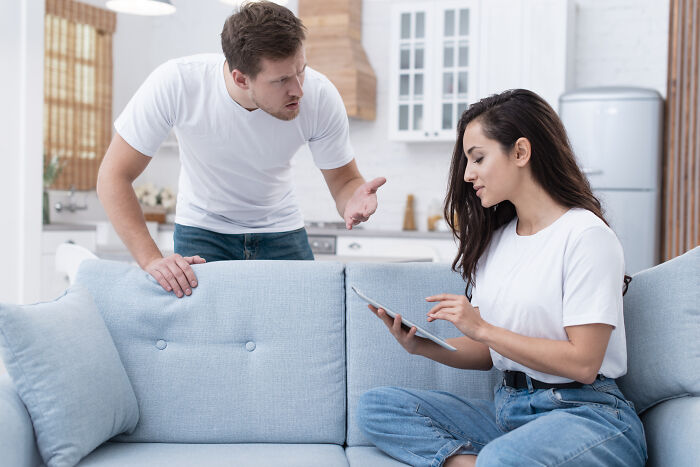 Man confronting woman on couch in modern living room, expressing anger about wife's infidelity and relationship issues.