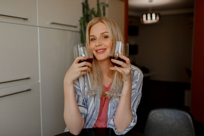 Young woman holding two glasses of red wine, smiling indoors, depicting a casual moment linked to wife’s infidelity themes.
