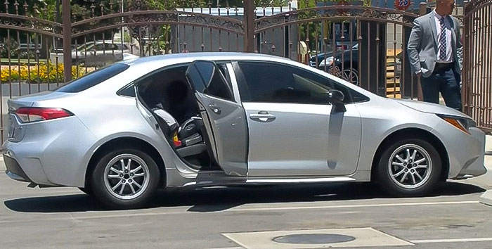 Silver car parked with rear door open, showing child car seat inside, while a man stands nearby by gated area. Silver car parked with rear door open, showing child car seat inside, while a man stands nearby by gated area.