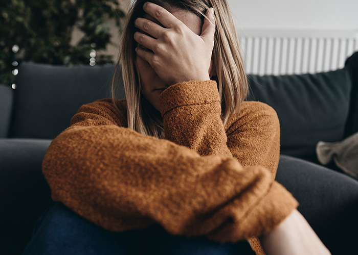 Woman in a brown sweater sitting on the floor covering her face, illustrating experiences of being gaslit by a doctor.