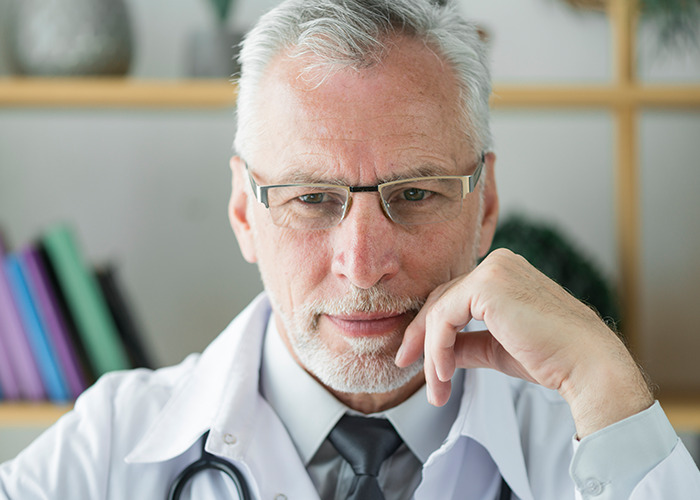 Mature male doctor wearing glasses and stethoscope looking thoughtful, representing gaslit by a doctor experiences.