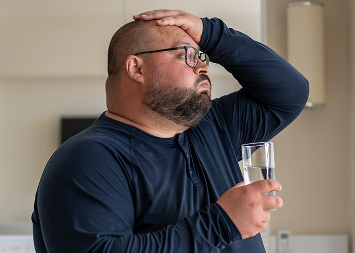 Man in glasses holding a glass of water, looking distressed and holding his head, illustrating being gaslit by a doctor.