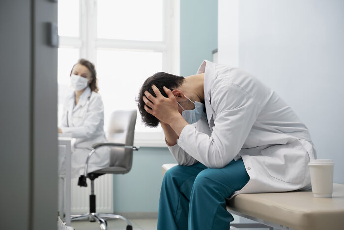 Medical professionals in a clinical setting, one stressed and holding his head, reflecting on dealing with dead bodies.
