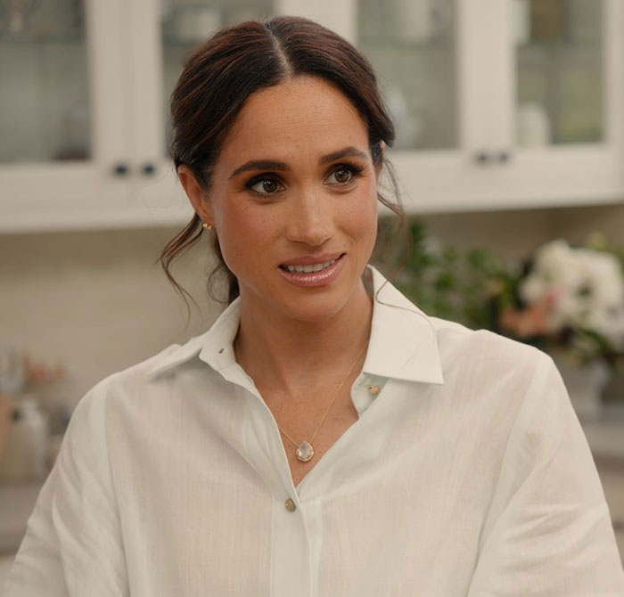 Meghan Markle in a white blouse indoors, reacting to accusations related to an embarrassing fruit-picking photo controversy.