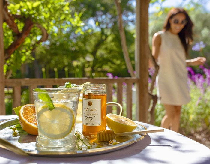 Close-up of a refreshing citrus drink and honey jar on a tray with a blurred woman in the background in a garden setting.