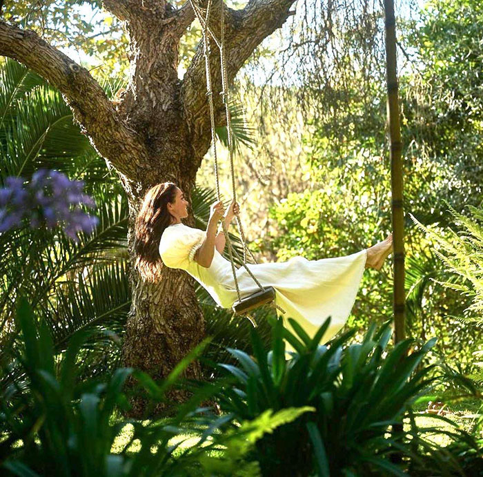 Meghan Markle on a swing in a garden setting, surrounded by greenery, linked to fruit-picking photo controversy.