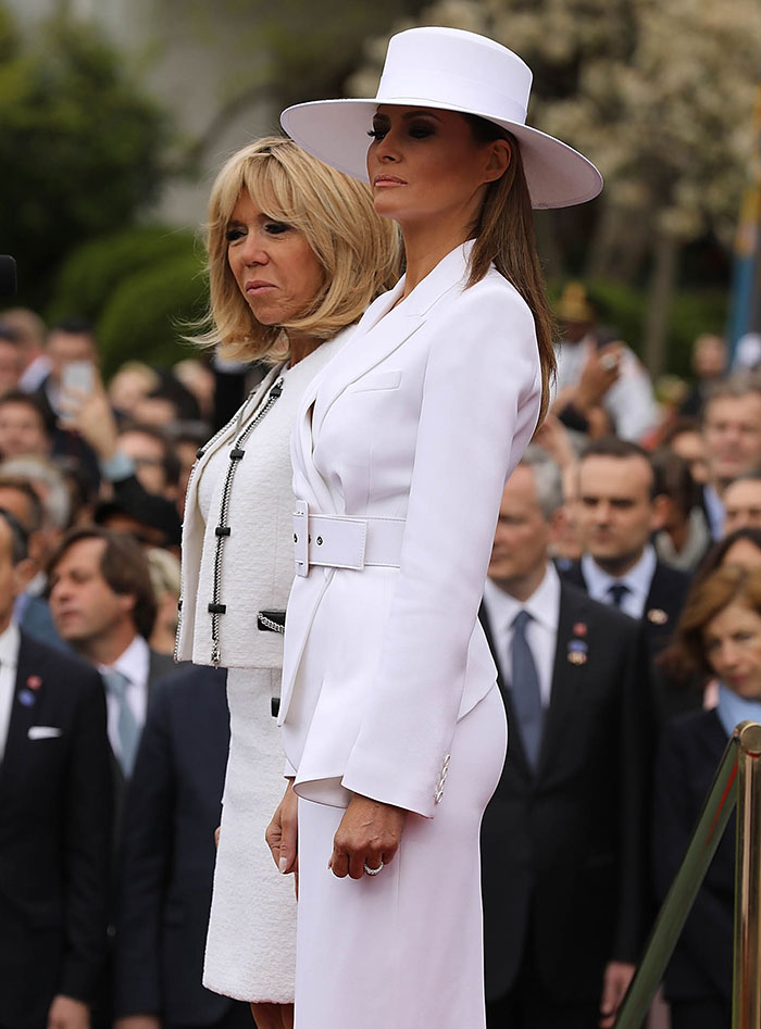 Melania Trump’s curves highlighted in white tailored outfit and hat, standing beside Brigitte Macron at public event. Melania Trump’s curves highlighted in white tailored outfit and hat, standing beside Brigitte Macron at public event.
