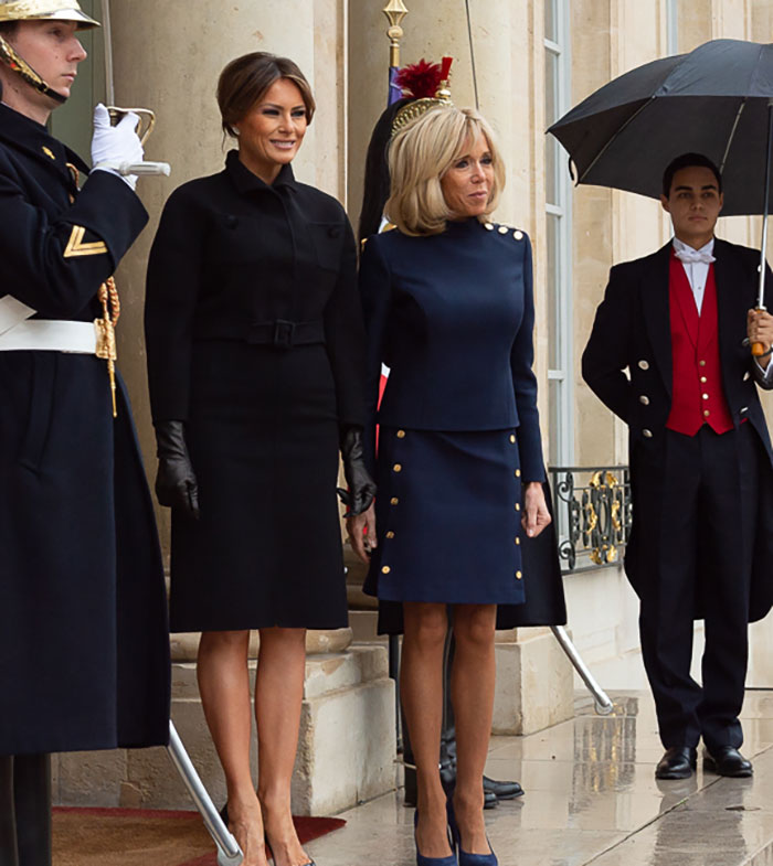 Melania Trump wearing a fitted black dress showcasing her curves next to Brigitte Macron outside a formal building. Melania Trump wearing a fitted black dress showcasing her curves next to Brigitte Macron outside a formal building.