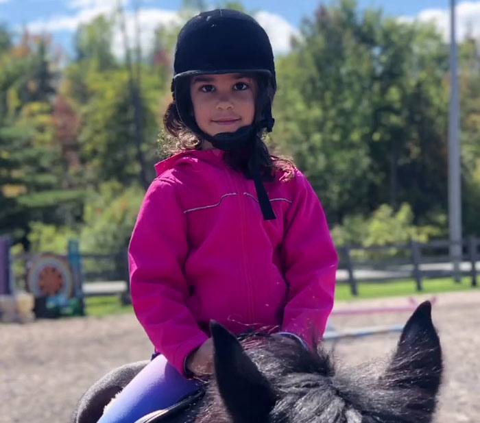 Young girl in pink jacket wearing helmet, sitting on a horse outdoors in a riding area with trees in background - 6