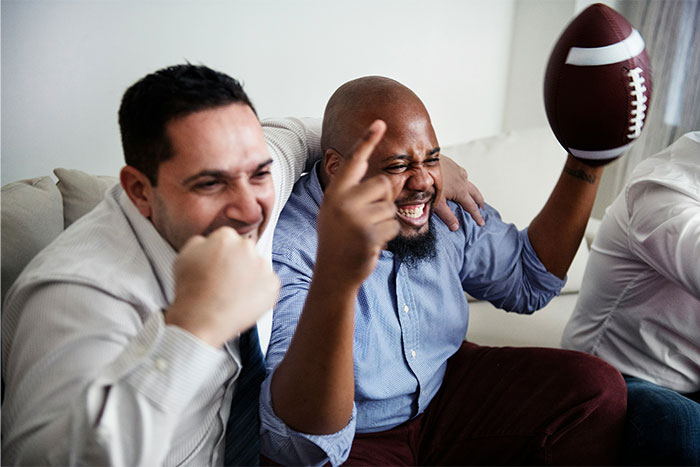 Two men celebrating excitedly on a couch while one holds a football, illustrating struggles men explain to women.