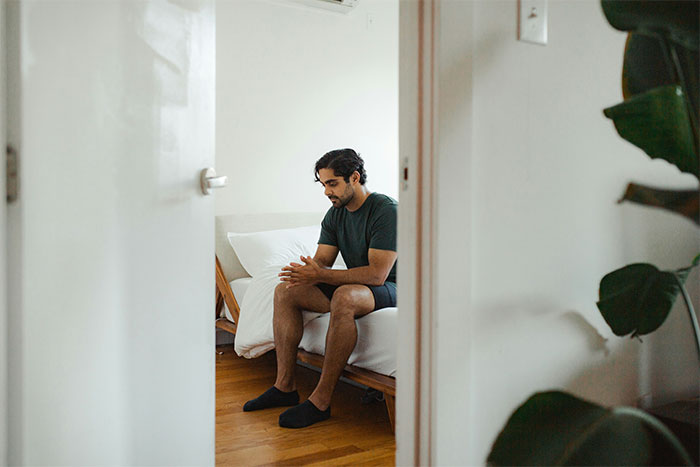Man sitting on bed with hands clasped, appearing thoughtful and frustrated, drives wife crazy concept.